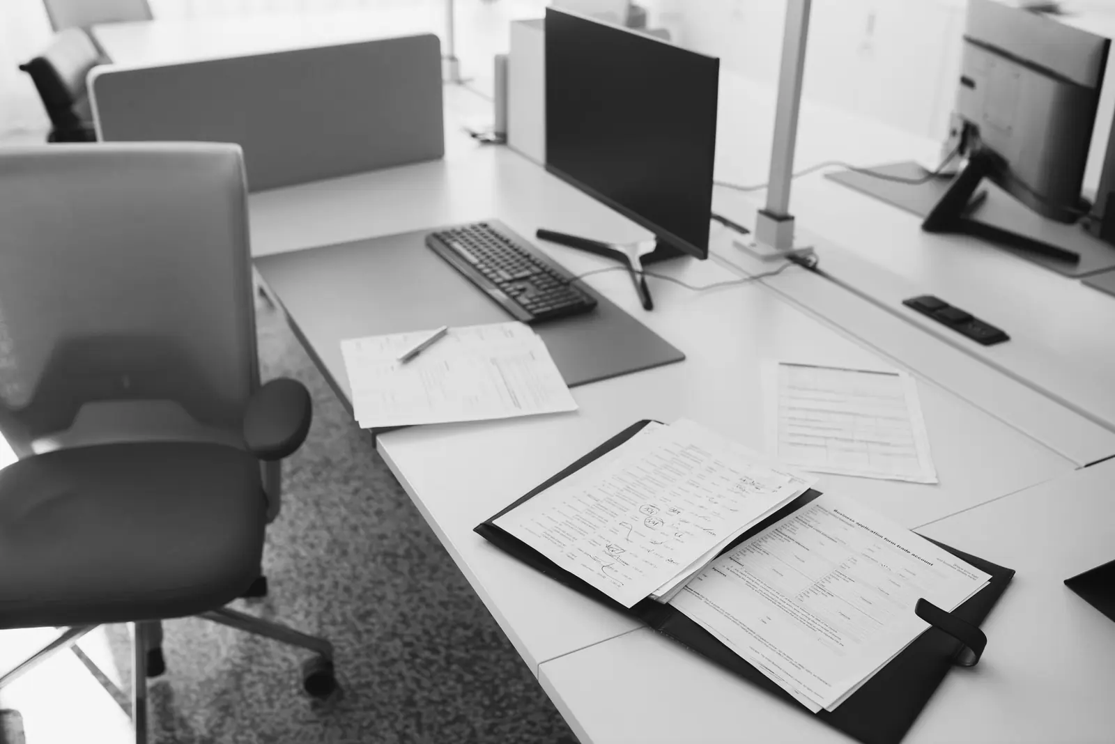 An empty office desk with a keyboard, a monitor and an open binder of printed forms beside a desk chair