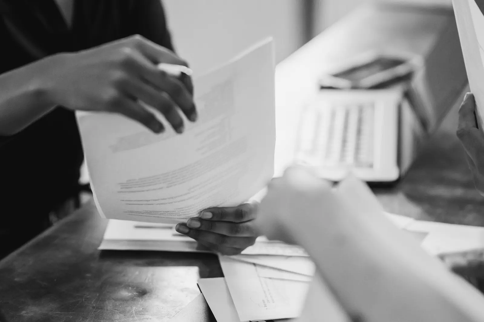 Close-up of hands passing printed pages across a desk, an open laptop out of focus behind