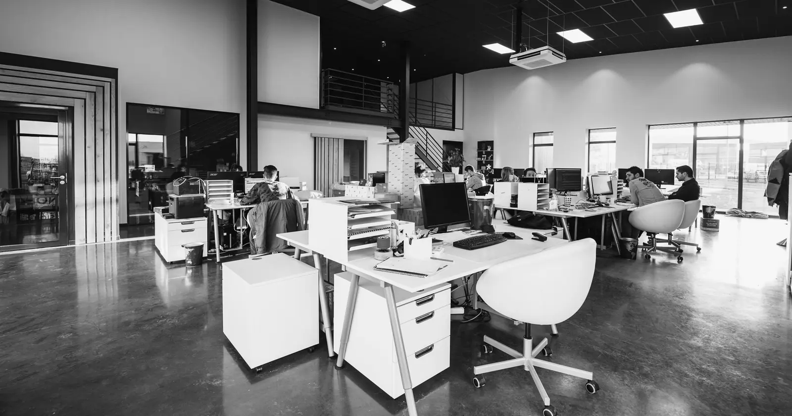 An open-plan office with white desks on a concrete floor, a few people working at the far desks